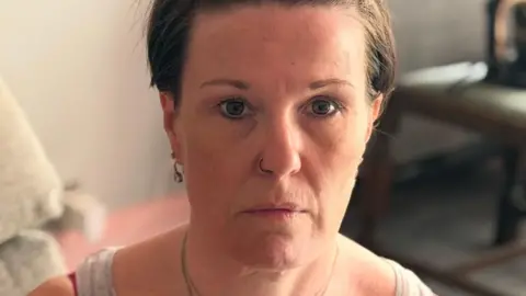 Lee-Ann Hawkes, who has short brown hair, sits on a sofa looking ahead with a serious expression. 