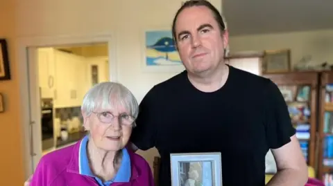 Tracey Miller / BBC A man wearing a black t-shirt and holding a framed photo of a woman. He has his arm around a woman wearing a purple and blue shirt who is standing to the right of him.