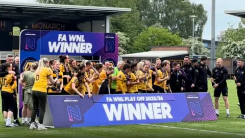 The team stand behind a ground-level placard that says 'Winners of FA WNL Play Off 2025/36'. Other team staff stand to the right hand side the women prepare to celebrate for photographs.
