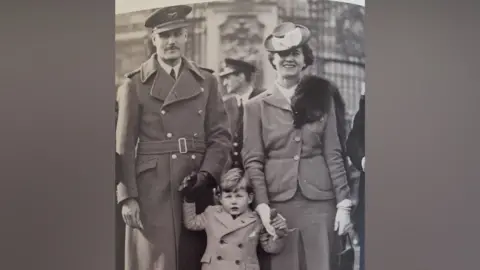 Norma Browne A black-and-white image from World War Two showing Group Captain Wilfred Surplice, his wife Cicely Wilson Surplice and their young son Roy. Wilfred is wearing a Royal Air Force uniform, including a belted greatcoat and cap with wings. A little boy, Roy, is in the middle, holding his parents' gloved hands, and he is wearing a short coat. Cicely is wearing a smart suit with a skirt, a hat tilted over her right eye and a fur tippet on one shoulder. 