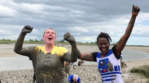 Lewis Adams/BBC BBC Essex presenters, Jodie Halford and Akylah Rodriguez cheer after making it through the mud. Jodie is particularly splattered.