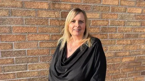 Pictured is a Paula Willcox with blonde shoulder length hair and she is wearing a black draped shirt with a silver necklace. She is standing and smiling against a pale brick wall.
