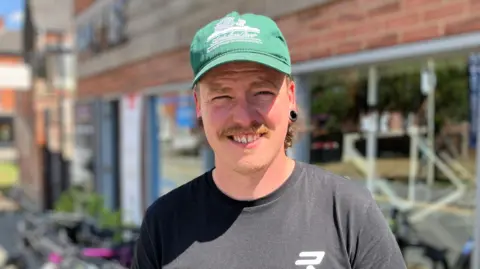Man wearing green baseball cap and black t-shirt smiling outside a cycle shop, which has large windows and a red brick exterior.
