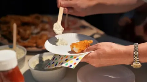 A breadcrumb fish finger on a paper plate. A hand is holding the plate and spooning a sauce on to the side. They are holding a paper napkin which has multicoloured spots.