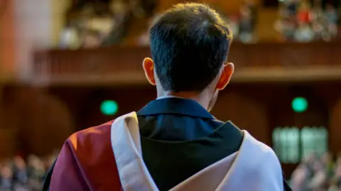 University of Bristol A view of Mr Fatthouh from the back. He has on a dark blue suit under his robes and is facing a crowd of more than 1,000 other graduates and their loved ones.