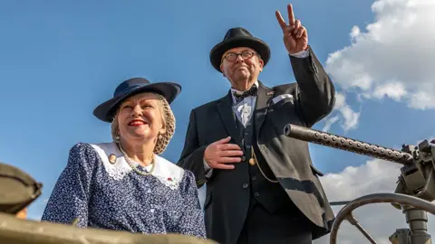 Andy Bell Images A woman in 1940s-style blue hat and dress with a big white collar on a military vehicle under a bright blue sky. A man stands to her side in a dark suit and bowler hat and holds up a V for victory sign. Part of a mounted military gun is visible beside them.