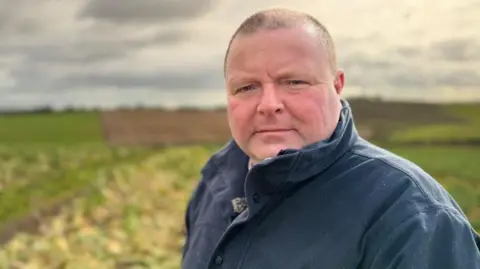 Stephen Murdoch is looking at the camera side on. He is wearing a navy coat and has very short brown hair.