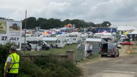 BBC A long distance shot of the show site. There are dozens of campervans and tents parked up to the left. hand side and a red and yellow circus top tent in the distance, as well as  a number of cars parked up along a section of fencing with festoon lights on it. A warden wearing a yellow high vis vest stands to the left of the image near a sign for the show.