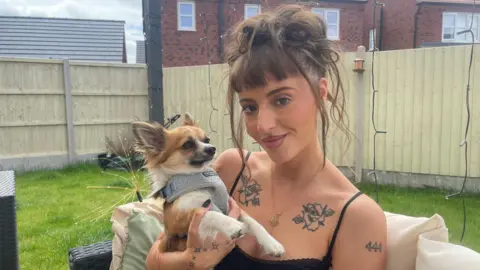 A young woman with dark hair worn up and a fringe sits in a garden holding a small tan and white long-haired chihuahua in a grey harness. She has floral tattoos across her chest and shoulders and the number 444 inked on one arm. Behind her are a wooden fence, a lawn and red-brick houses.