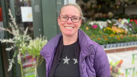 A woman smiling outside a flower shop. She is wearing purple frame glasses. a purple quilted jacket and black t-shirt with white stars printed on it.