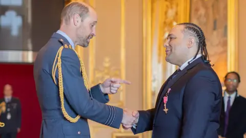 The Diana Award Alex Holmes shaking the hand of Prince William, the Prince of Wales, in a room in Windsor Castle. Prince William is wearing an army uniform, and is pointing at Alex. Alex is wearing a blue suit, with his OBE medal on his lapel. His hair is braided. 