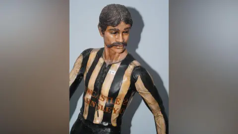 Anderson & Garland The life-sized Newcastle United figure which has brown, short hair and a large brown moustache. The figure is looking to the right with its arms slightly outstretched. It is wearing a long-sleeved black and white shirt which has Newcastle 2 Barnsley 0 written on the front in red letters. It is in front of a blue background with a shadow behind it to the right.