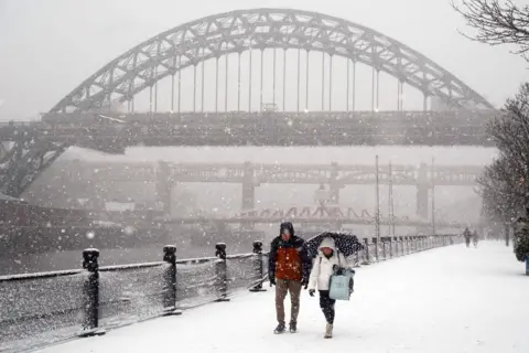 PA Media The Tyne Bridge is slightly obscured by the falling snow with the Swing Bridge and High Level Bridge beyond. Two people, wrapped up in coats and one with a black and white spotty umbrella, are walking along the snow-covered quayside. Black bollards with railings run alongside them, with the River Tyne to their right.