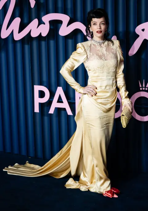 Getty Images Lily Allen wearing a gold dress with red shoes and a matching gold clutch, with her dark hair curled and tied back