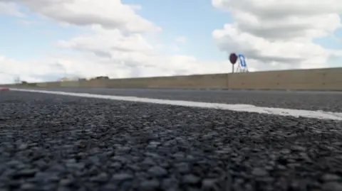 A close-up low angle photo of a tarmaced road. There is a white line and on the far side of the road a low wooden barrier. Overhead the sky is blue with white clouds.