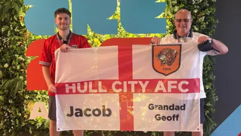 A grandad and grandson wearing England football shirts and holding a white and red flag of St George in front of a floral background. On the left, a young man with brown hair and a closely cropped beard wearing a red shirt. On the right, an older man wearing sunglasses and a white shirt. The flag has the words "Hull City AFC written across the centre, the names "Jacob" and "Grandad Geoff" in the lower quadrants and a Hull City tiger badge in the upper right quadrant. 