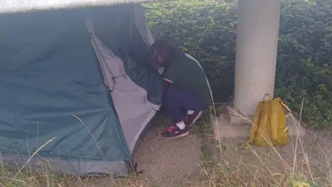 Maggs Day Centre A woman with dark hair squats outside a tent and looks inside. A yellow backpack is on the ground near a pillar.