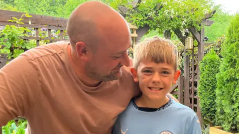 Mike, who wears a light pink shirt, smiles at James. He has his arms around his shoulders. James wears a light blue Manchester City shirt and has blonde hair, and smiles directly at the camera.