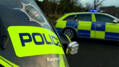 BBC police service of northern ireland creast on front of a police motrike which is parked in front of a police car