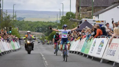 George Ledger/Project Genesis A female cyclist throws her arms in the air as she crosses the finishing line of the race on 22 June. Another cyclist and a man on a motorbike are following behind. Spectators are lining both sides of the street.
