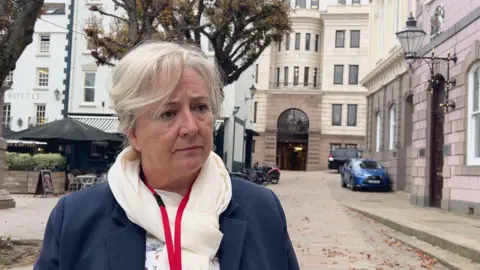 Deputy Karen Wilson is frowning slightly. She is standing in a street with buildings behind and to the side of her. She is wearing a navy jacket, a cream scarf and a red lanyard.