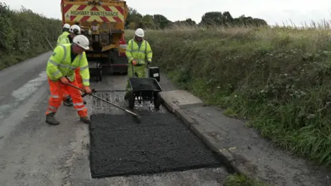 Workers in hi-vis are carrying out work on a road. They appear to be fixing potholes . The four workers are wearing hard hats and are standing in a country lane next to a lorry. 