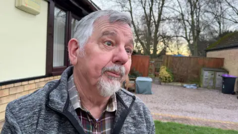 A man with short grey hair and a grey goatee beard looks off camera as he stands in the garden of a home. He is wearing a grey fleece and dark checked shirt. A brick building with cream cladding stands behind him. A yard covered in pebbles can be seen, stretching towards a brown wooden fence and trees.
