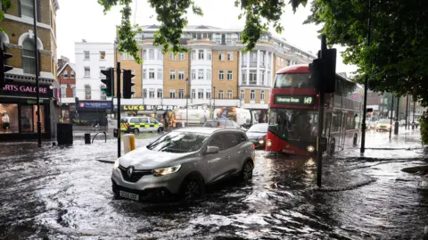 Leon Neal/Getty Images A flooded London street, with a silver car and red London bus driving through the water at a crossing. There are flats and shops visible in the background and a tree hangs over the street, visible in the top right of the picture