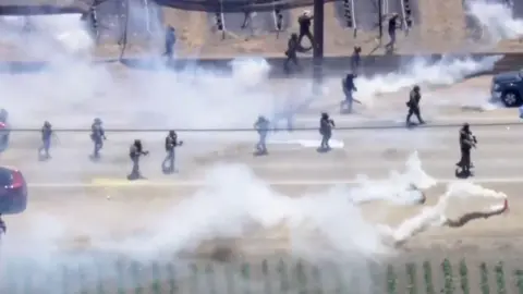 Aerial view of immigration agents deploying tear gas on protesters on a road.