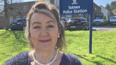 Joanna Brittan looking at the camera and smiling. She has a short dark bob and is wearing a pearl necklace and earrings. She also has on a purple top. She is pictured from the shoulders up. There is a grass back behind her with a sign on it saying "Police Station".