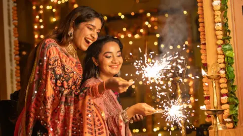 Two women are holding sparklers. They have dark brown hair and are wearing orange and pink dresses with large gold jewellery. Around them are string lights and golden candle holders