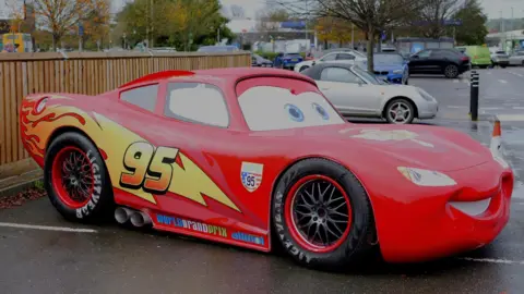 Luke Russell/Dead Sharp Media A bright red car, which is a model of the character Lightning McQueen from the Disney Pixar film Cars. It is parked in a car park with other cars in the background.