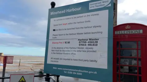 A sign depicting the fees at the harbour. A beach can be seen in the distance, with the sea on the horizon.