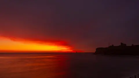 Looking out to sea at Tynemouth. The sky is largely dark but to the left there is a bright orange and red patch which looks to be spreading further right. The priory is to the right of the photo and is completely in shadow. In the distance the pier and lighthouse can be seen.