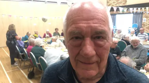 Ivy's son Barry stands in front of tables with guests at party 