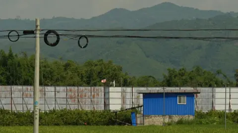 Xiqing Wang/BBC  A high, metal border fence cuts through fields between China and Myanmar in Ruili 