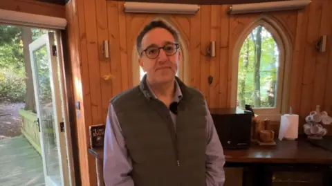 Dr Yaz Romahi wearing a purple shirt and a green gilet. He has short brown hair and glasses. He is stood in front of a treehouse wall, which is wooden.