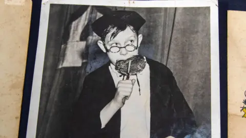 Hansons A young Eric with glasses, sucks on a large lollipop, wearing a school hat and cloak