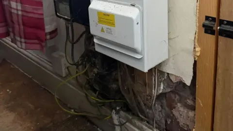 Electric meter fixed to wall with exposed live wiring hanging down underneath and shoddy plasterwork.
