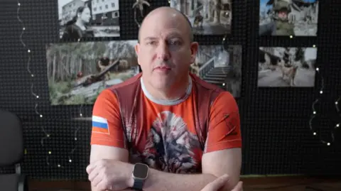 X A bald man looking at the camera wearing a t-shirt with a wolf on the front and a Russian flag on the sleeve