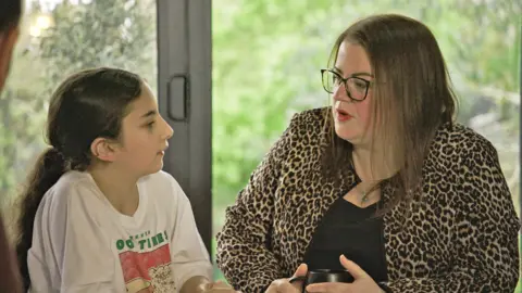 A girl with long dark hair and a white T-shirt with a pink and green print on the front looks towards a woman with brown hair, glasses and wearing a leopard-print jacket. A garden - through glass in the background - is out of focus.