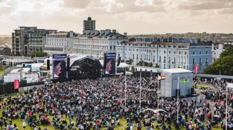 Summer Sessions Aerial view of a large crowd at summer sessions in Plymouth. Large crowds have gathered on the Hoe to watch artists on a large stage. Residential properties are located behind the stage. 