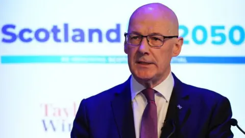 John Swinney, who is bald and is wearing glasses, is wearing a blue suit, white shirt and purple tie and is in front of a background saying Scotland 2050. He has a saltire tie-pin on his left lapel.