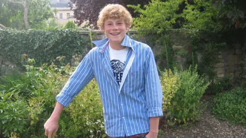 Handout A teenage boy with curly blond hair smiles as he stands in a garden. There are bushes behind him and he is propping himself up on something out of shot, perhaps a shovel. He is wearing a blue striped shirt which is unbuttoned to show a white T-shirt underneath. There is a big house in the distance.
