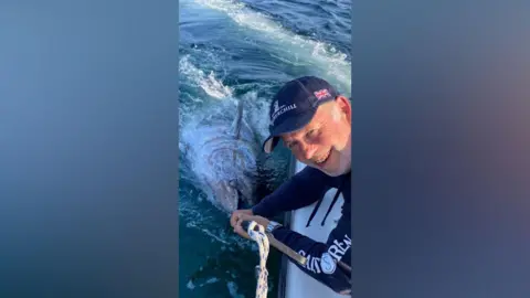 Rupert Drury A middle-aged man in a blue baseball cap smiling with a large fish hooked on to a pole in his arms. The man is leaning over a boat in open waters.
