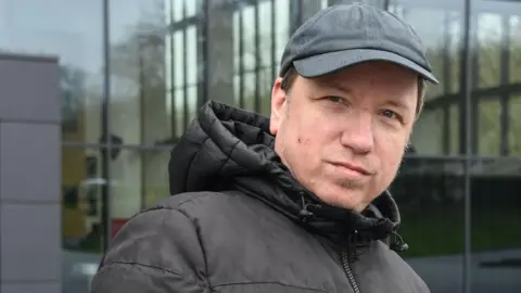 Dirk Hähnel looks at the camera. He is wearing a black jacket and black hat