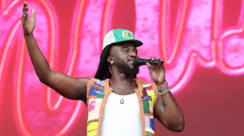 EPA A man wearing a colourful crochet vest is on stage in front of a pink and red background. He is holding one of his arms into the air.