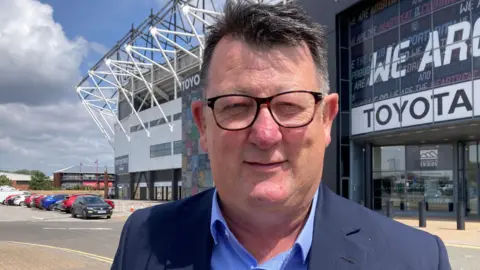 Paul Peacock outside Pride Park. He is smiling and is wearing glasses, a shirt and a jacket. 