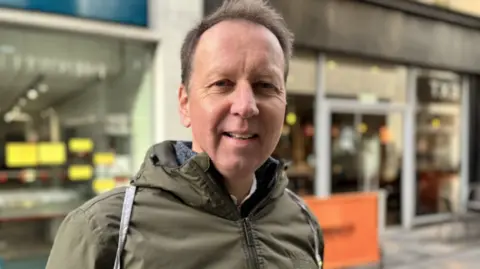 Philip Kyle has short dark hair and is wearing a khaki green coat with a hood and is standing in a high street, with shop windows behind him.