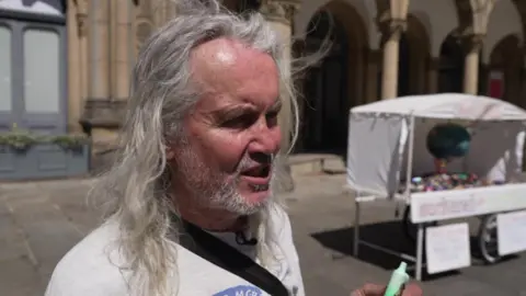 A man with long grey hair holds up a green vape. Behind him is a vape sculpture 